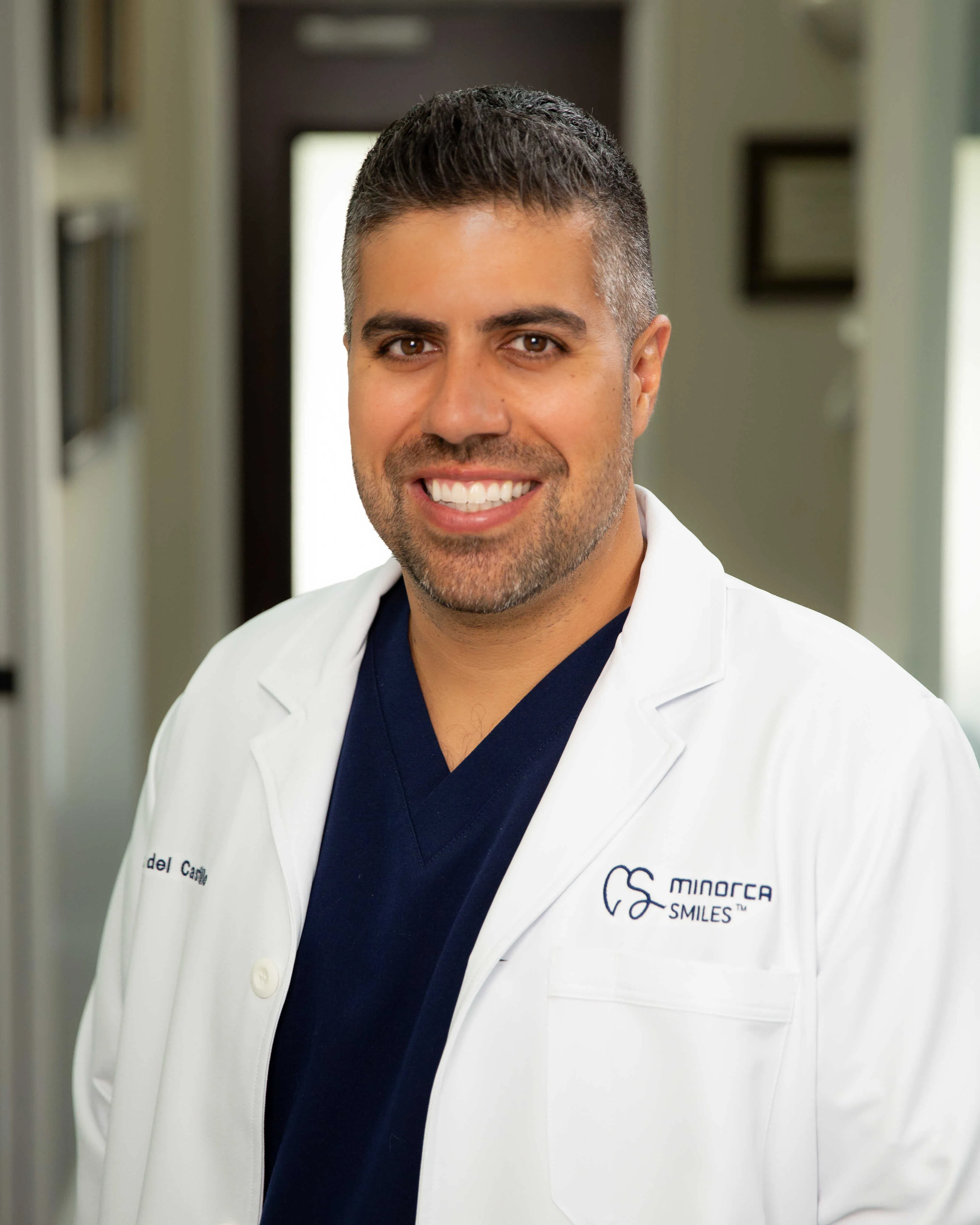 Doctor Raymond Del Castillo portrait wearing blue scrubs and a white Minorca Smiles lab coat