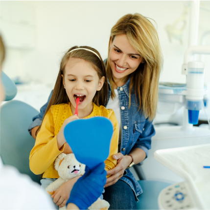 Smiling young girl holding a mirror and teddy bear during dental visit with her mother beside her.