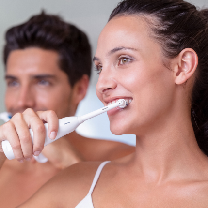 Smiling woman using an electric toothbrush with a man brushing in the background.