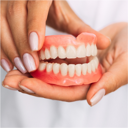 Hands holding a full set of removable dentures with natural-looking teeth and pink gums.
